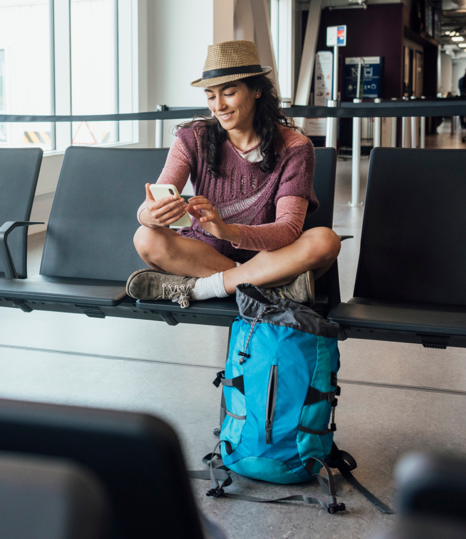 Young woman sitting with crossed legs looking at her phone at the airport with travel insurance for missed connections
