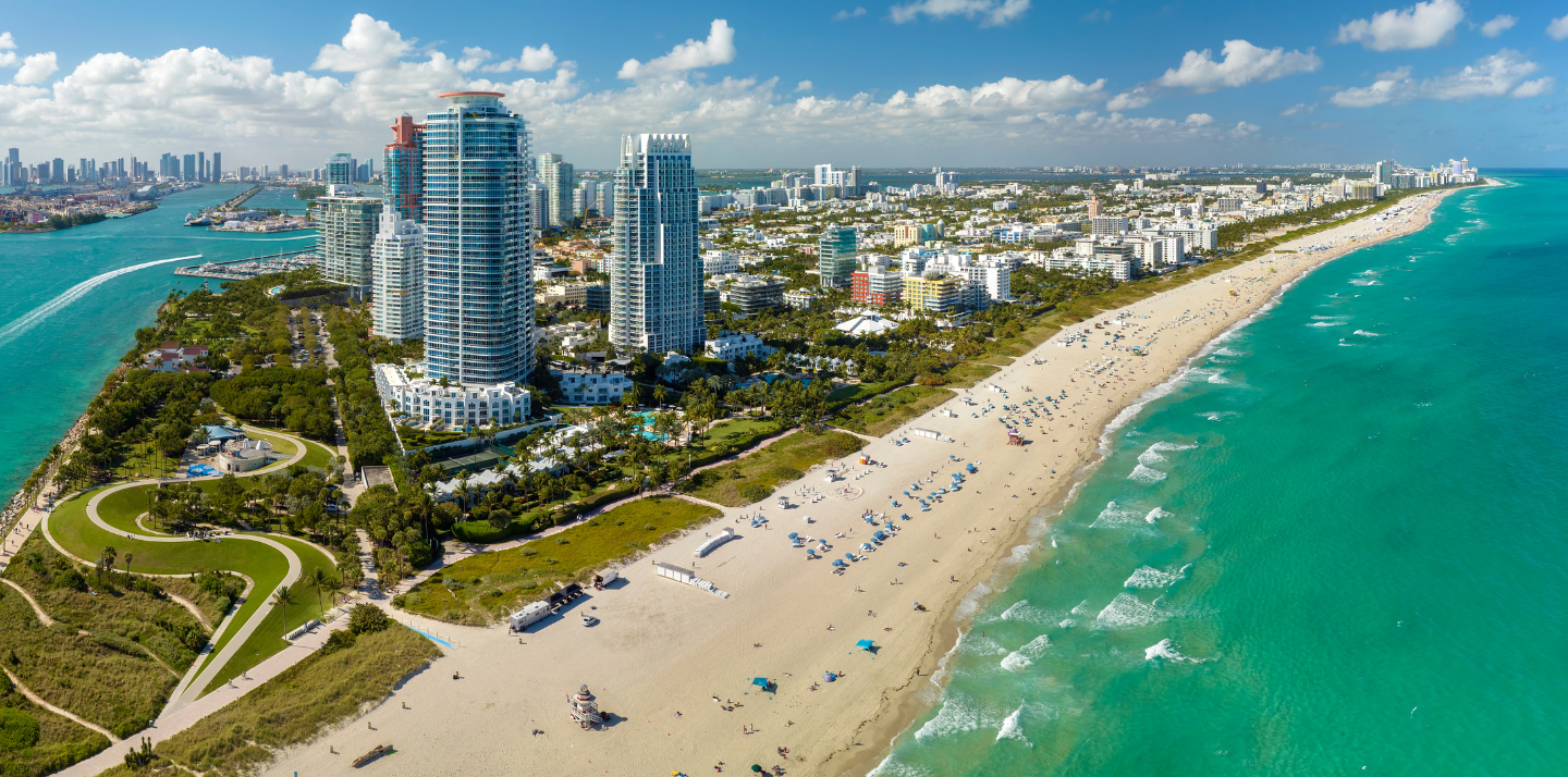 Aerial view of Miami Beach featuring the coastline with sunbathers on the sand and the city skyline in the background.