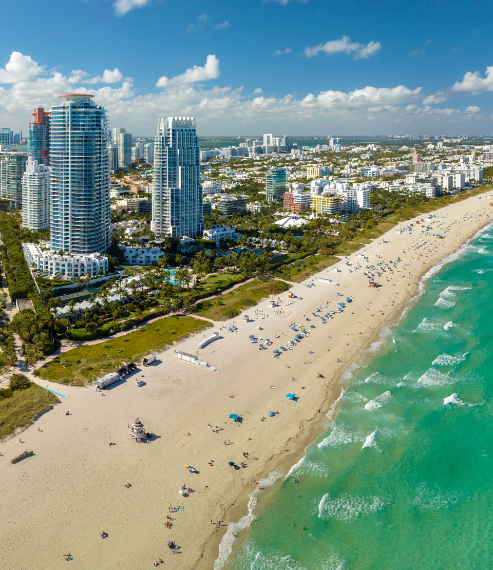Aerial view of Miami Beach featuring the coastline with sunbathers on the sand and the city skyline in the background.