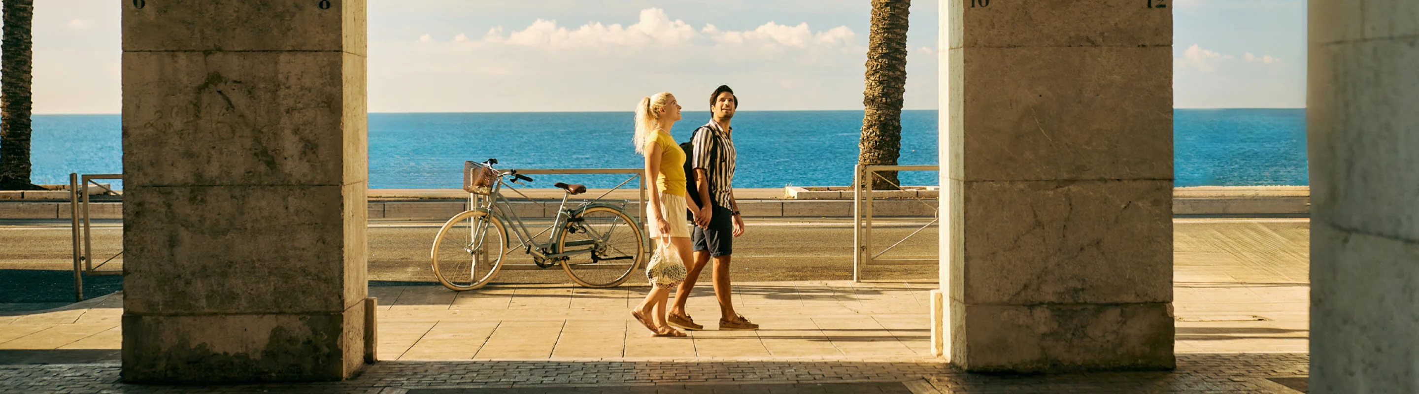 a young couple with travel insurance for beach trips holds hands as they walk palm trees and the ocean 