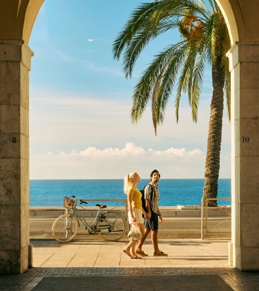 a young couple with travel insurance for beach trips holds hands as they walk palm trees and the ocean 