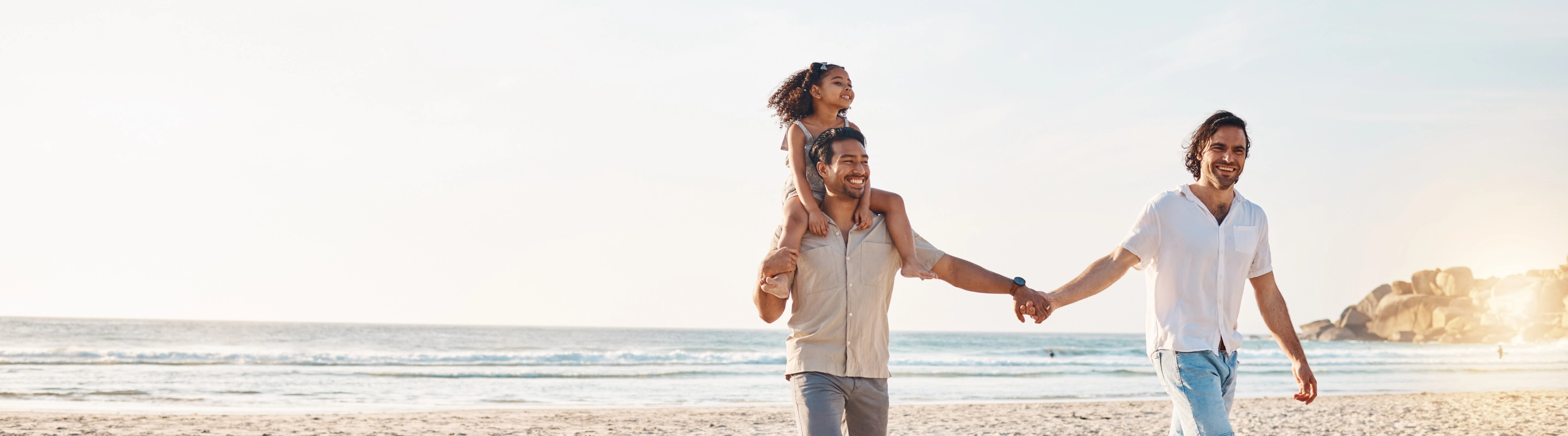 a couple with LGBTQ+ travel insurance hold hands on a beach, their daughter riding on one of their shoulders