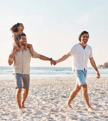 a couple with LGBTQ+ travel insurance hold hands on a beach, their daughter riding on one of their shoulders