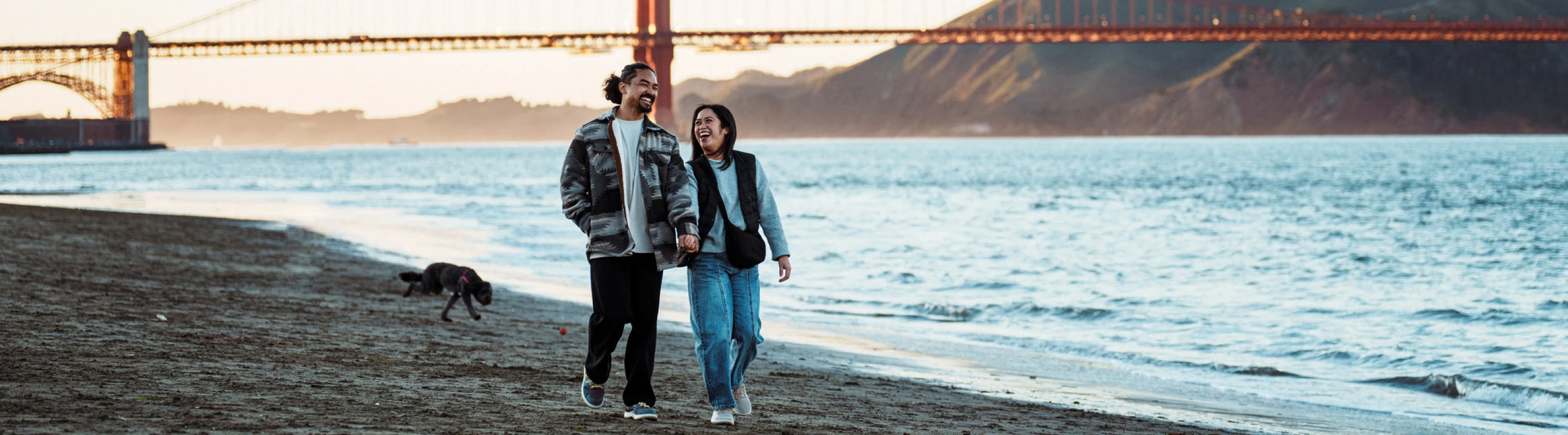 a couple with domestic travel insurance walk along San Francisco's shoreline with the Golden Gate Bridge in the background