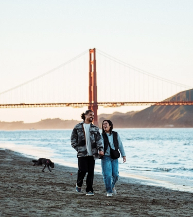 a couple with domestic travel insurance walk along San Francisco's shoreline with the Golden Gate Bridge in the background