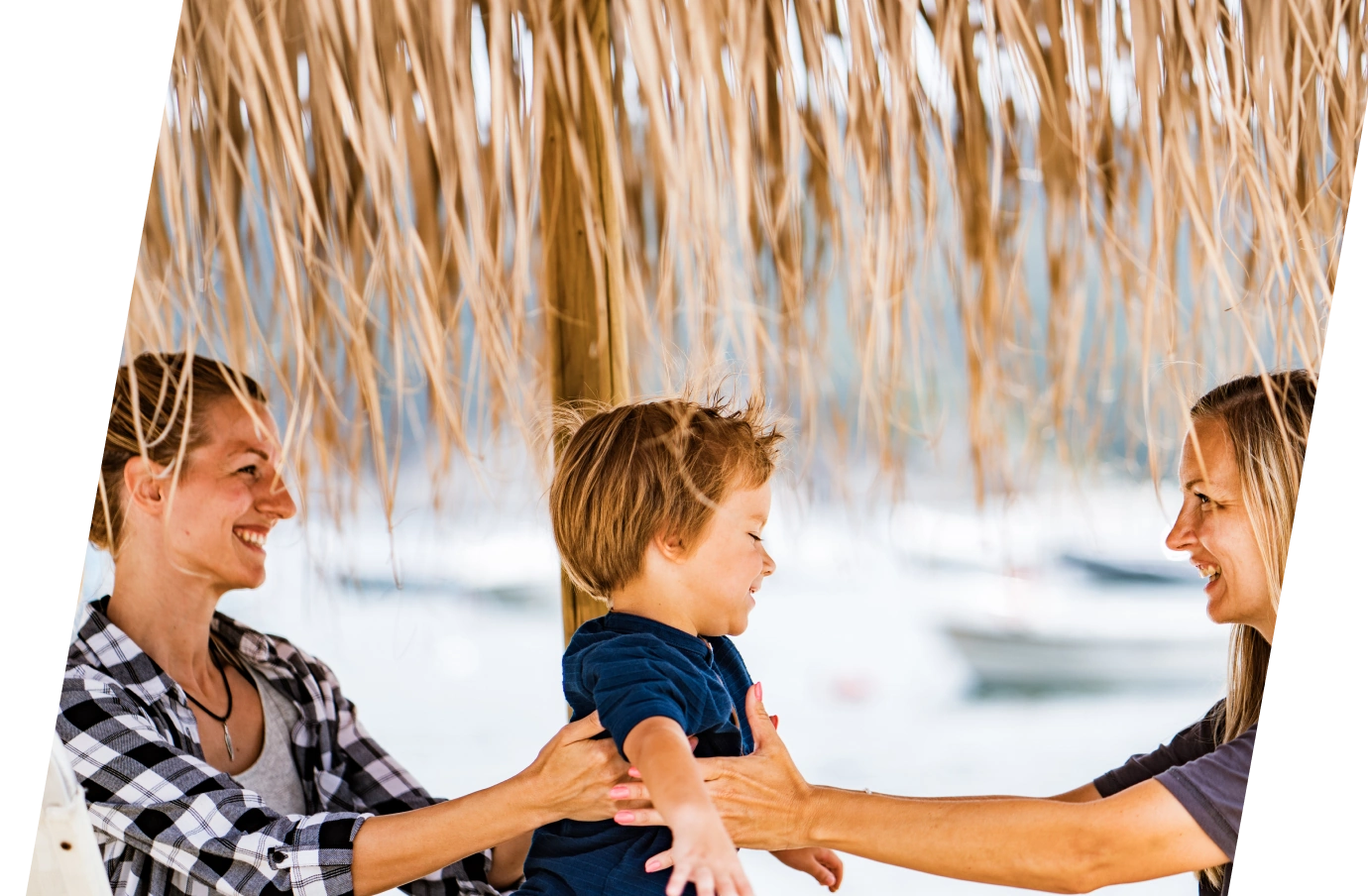 a young couple with LGBTQ+ travel insurance hold their toddler son up in the air under a thatched umbrella 