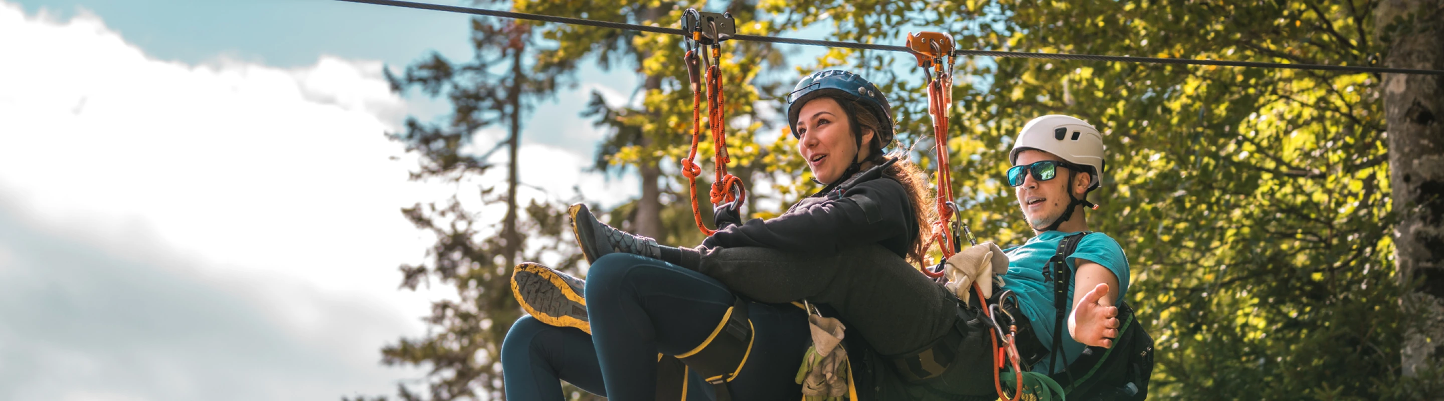 a young couple with adventure travel insurance tandem zip line through treetops on a blue-sky day