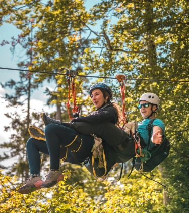 a young couple with adventure travel insurance tandem zip line through treetops on a blue-sky day