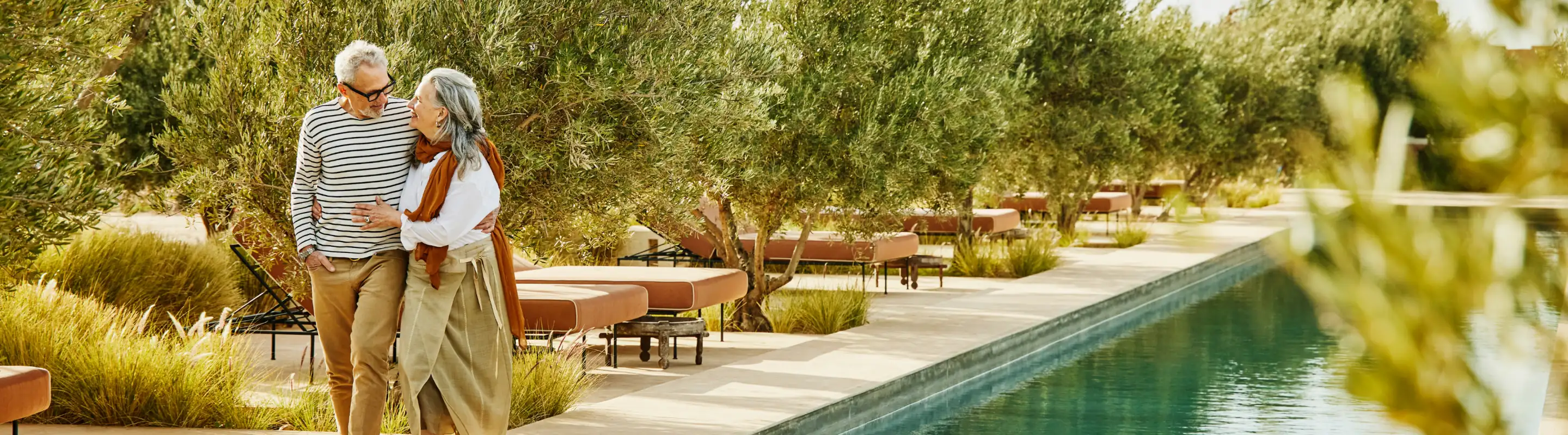 an elderly couple with long-term travel insurance walks through a garden-lined poolside resort 