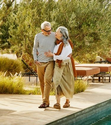 an elderly couple with long-term travel insurance walks through a garden-lined poolside resort 