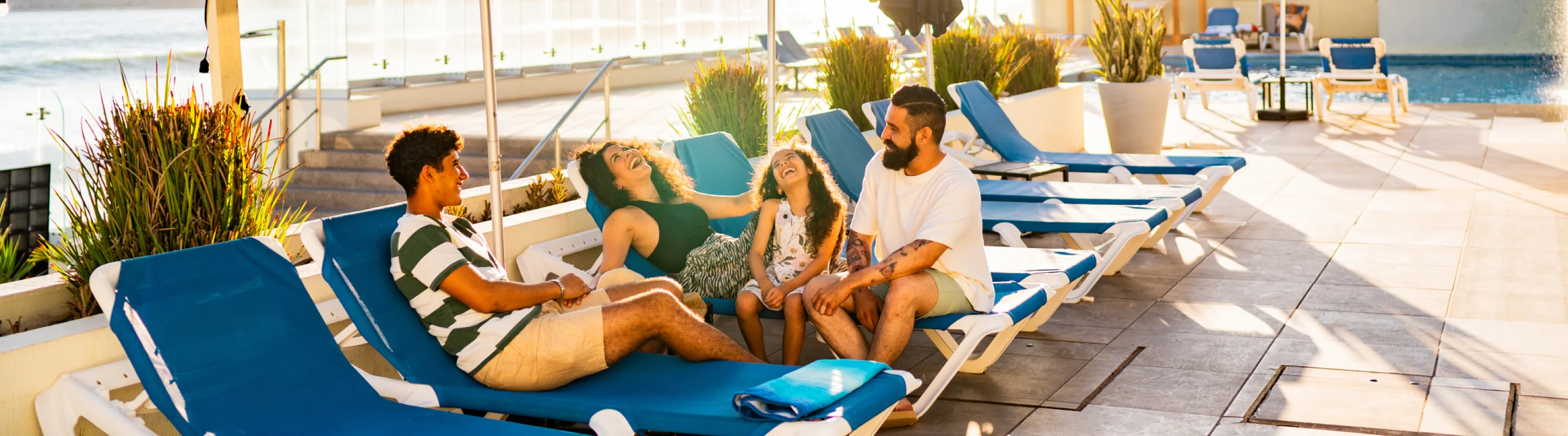 Parents with travel insurance for Mexico laughing with their teen kids on lounge chairs by the pool with the ocean in the background