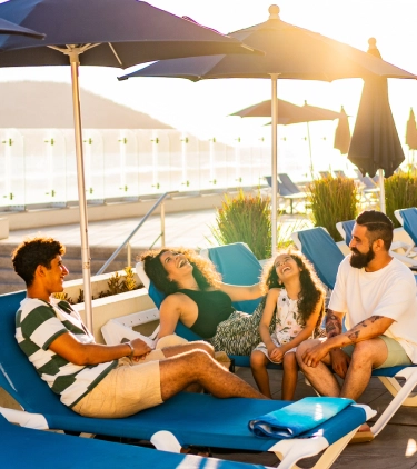 Parents with travel insurance for Mexico laughing with their teen kids on lounge chairs by the pool with the ocean in the background