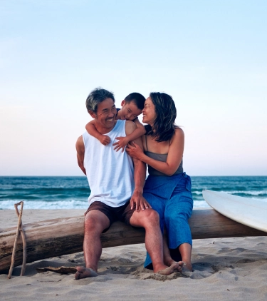 parents with inexpensive travel insurance sitting on a log on the beach with their child at sunset