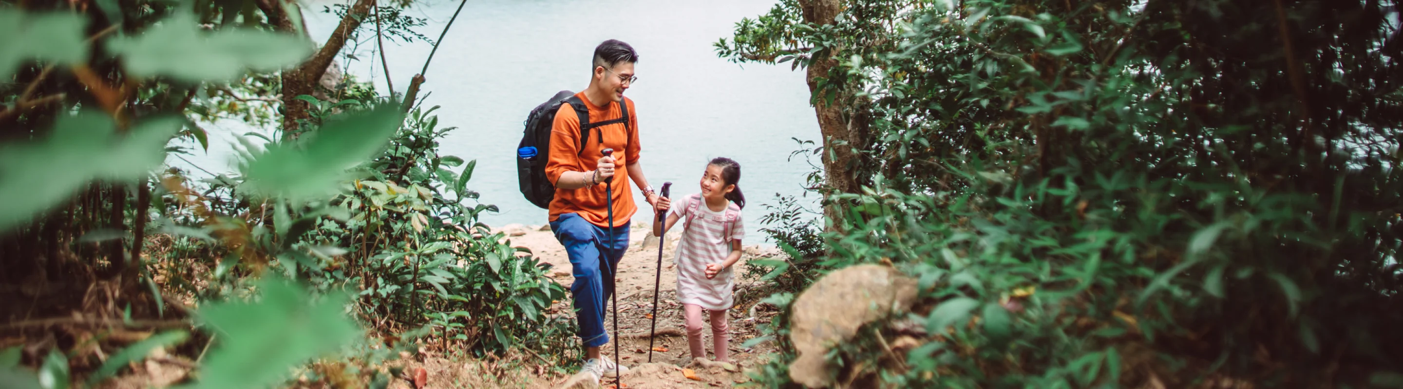 Father with comprehensive travel insurance smiling at his daughter while hiking in the woods with a lake in the background