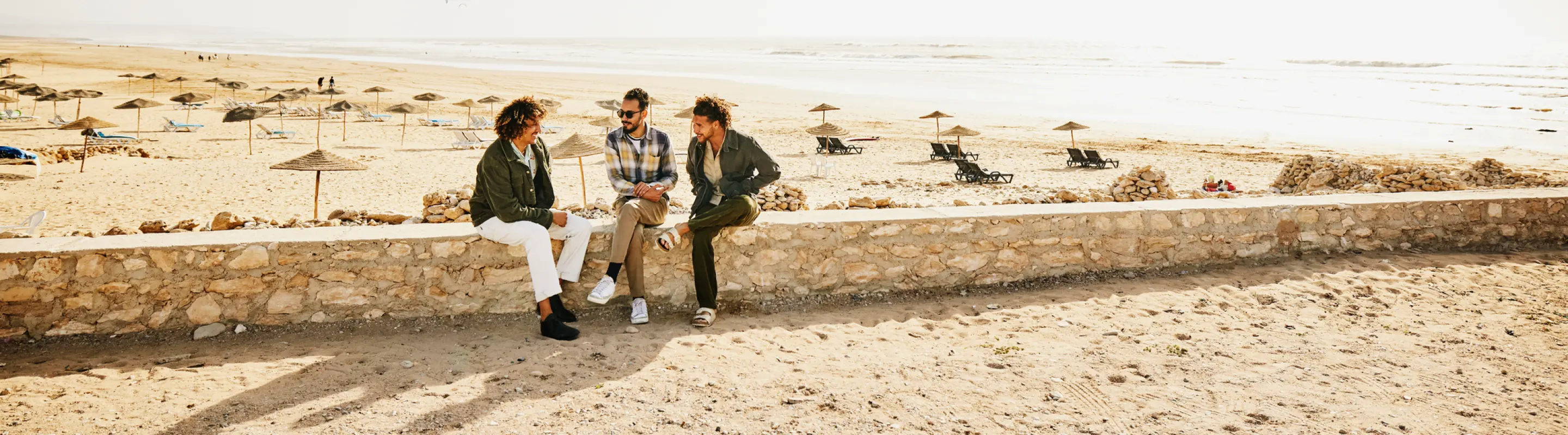 Three adults with last-minute travel insurance sitting on a stone wall and talking with the beach in the background