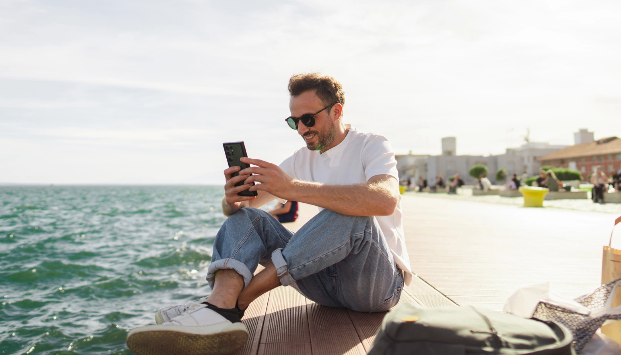 a young man sits by the ocean while he researches the difference between a travel alert and advisory on his phone
