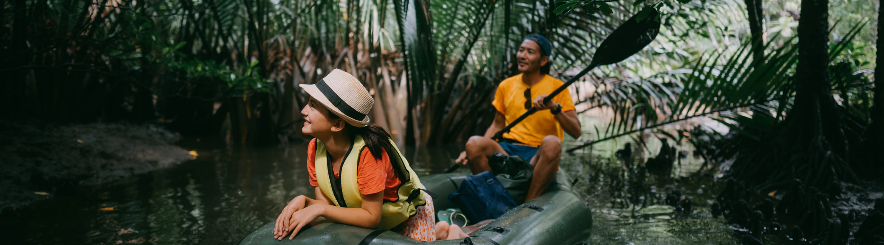 a man and woman with international travel insurance share a kayak and paddle through a mangrove with palm fronds