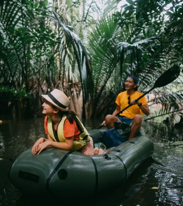 a man and woman with international travel insurance share a kayak and paddle through a mangrove with palm fronds