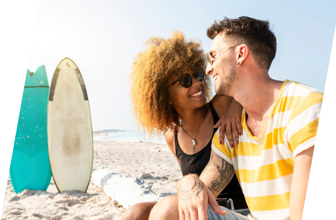 a young, mixed-race couple with travel insurance for beach trips sits on the sand by the ocean with their surfboards nearby