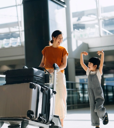 a mother with domestic travel insurance pushes a luggage cart in an airport while her daughter walks beside her