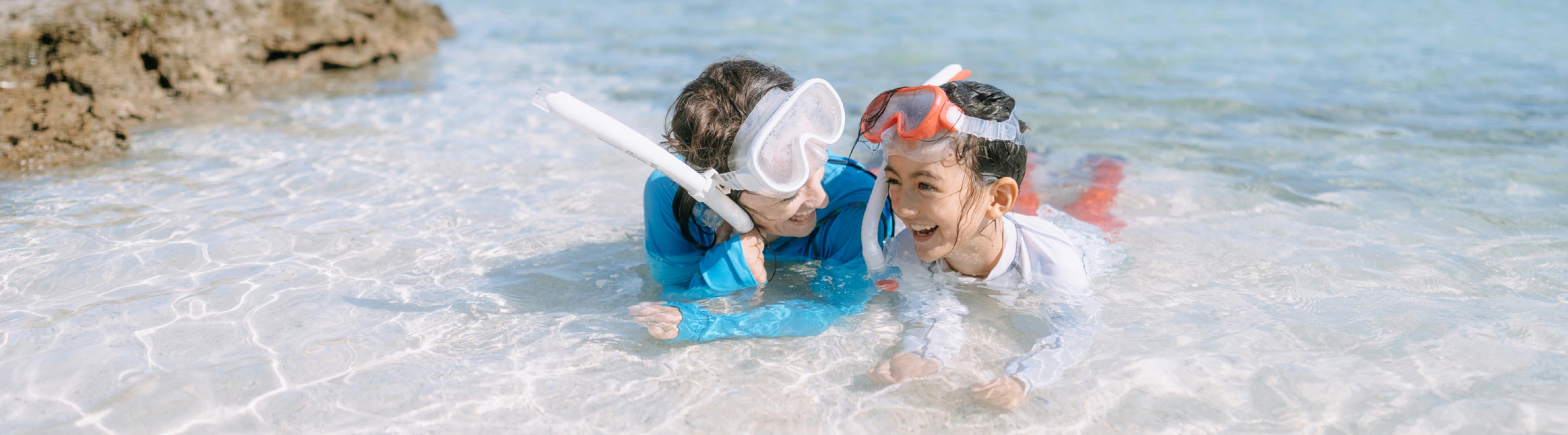 a mother-daughter duo with adventure travel insurance lay mid-shore in crystal clear water with snorkel gear on their heads