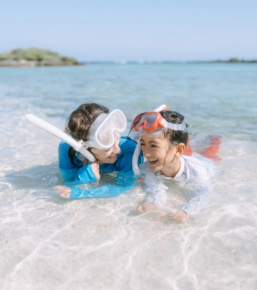 a mother-daughter duo with adventure travel insurance lay mid-shore in crystal clear water with snorkel gear on their heads
