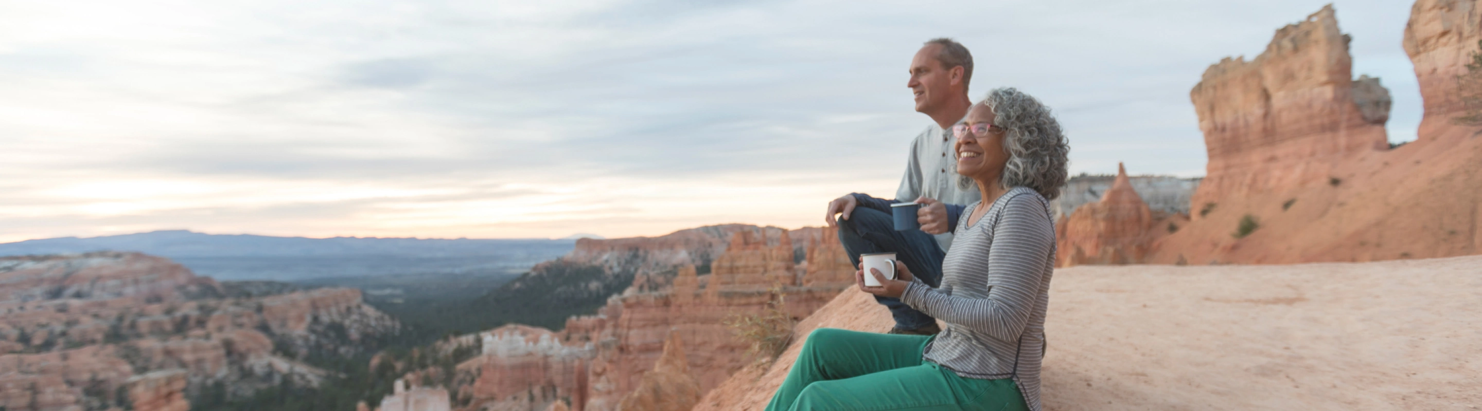 an older couple with domestic travel insurance sits on a red rock canyon's ledge in Utah and soaks up the lookout's views