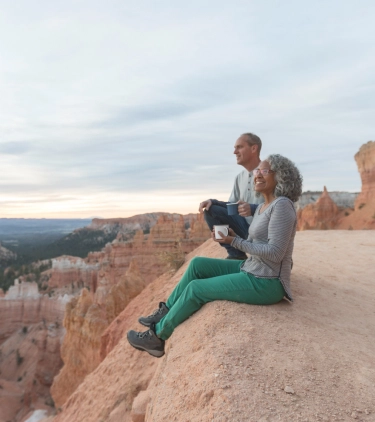 an older couple with domestic travel insurance sits on a red rock canyon's ledge in Utah and soaks up the lookout's views