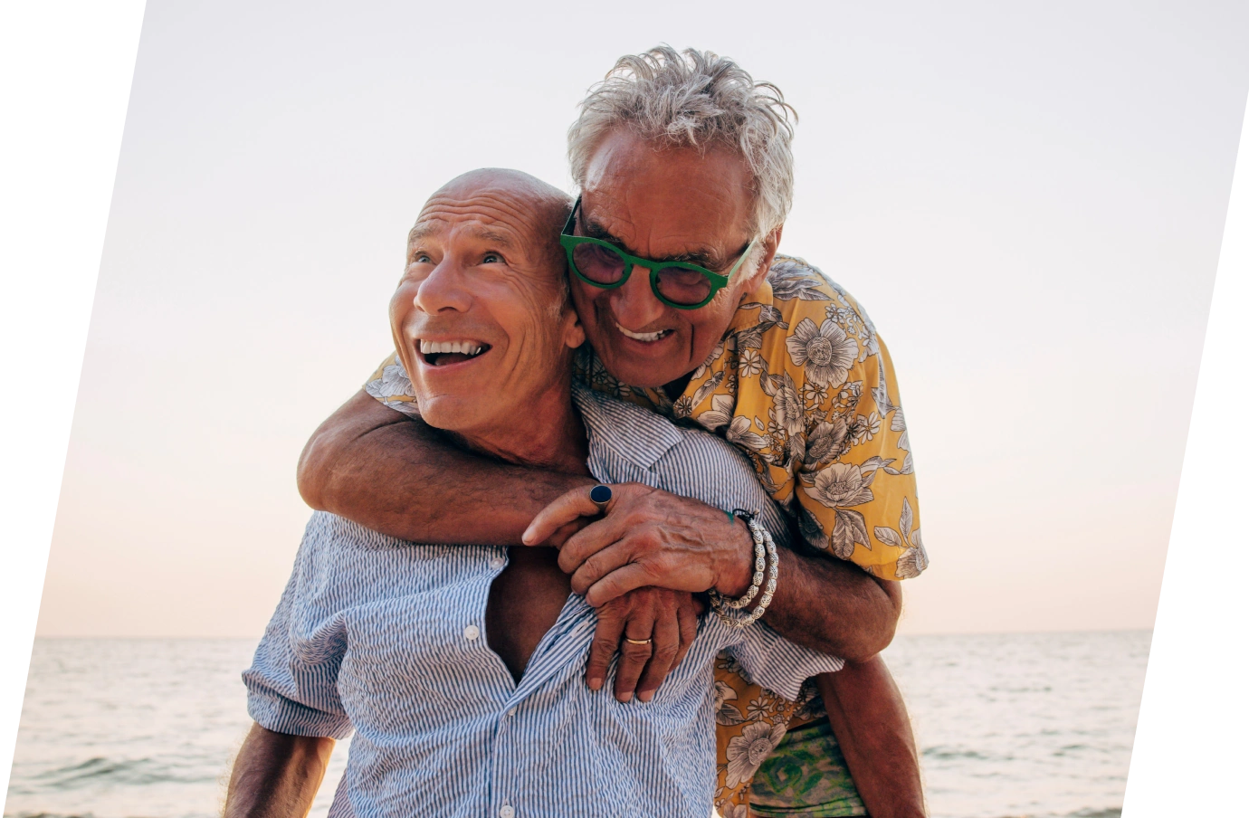 an older couple with LGBTQ+ travel insurance smiles and embraces one another in a seaside setting 