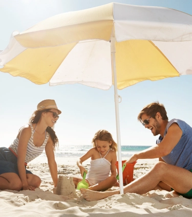 parents with travel insurance for beach trips help their young daughter build a sandcastle by the ocean on a sunny day