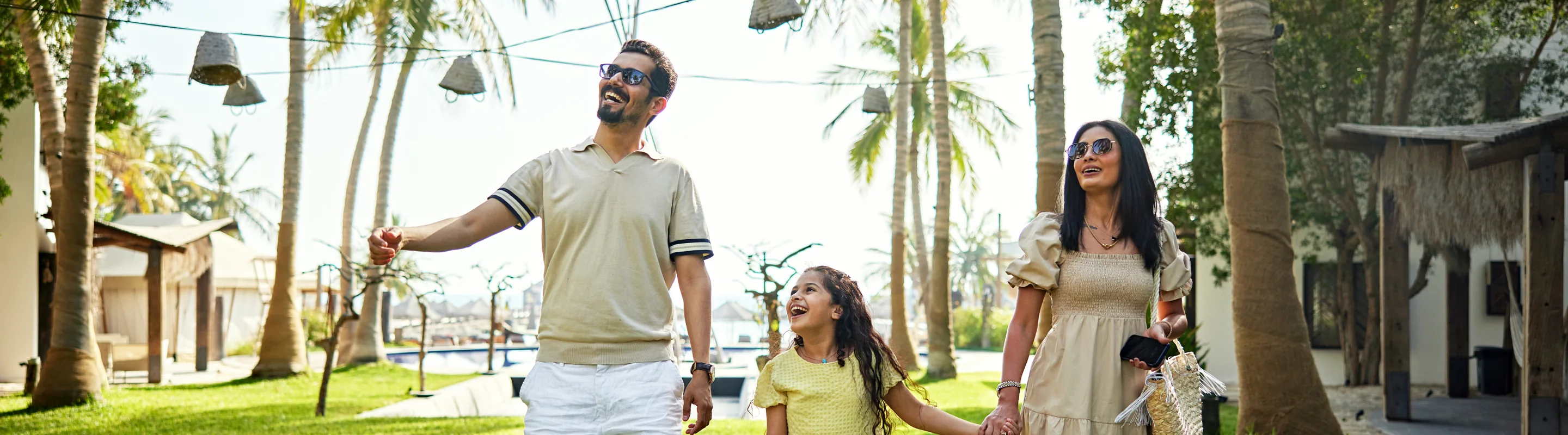 two young parents with long-term travel insurance walk alongside their daughter through a tropical resort with palm trees