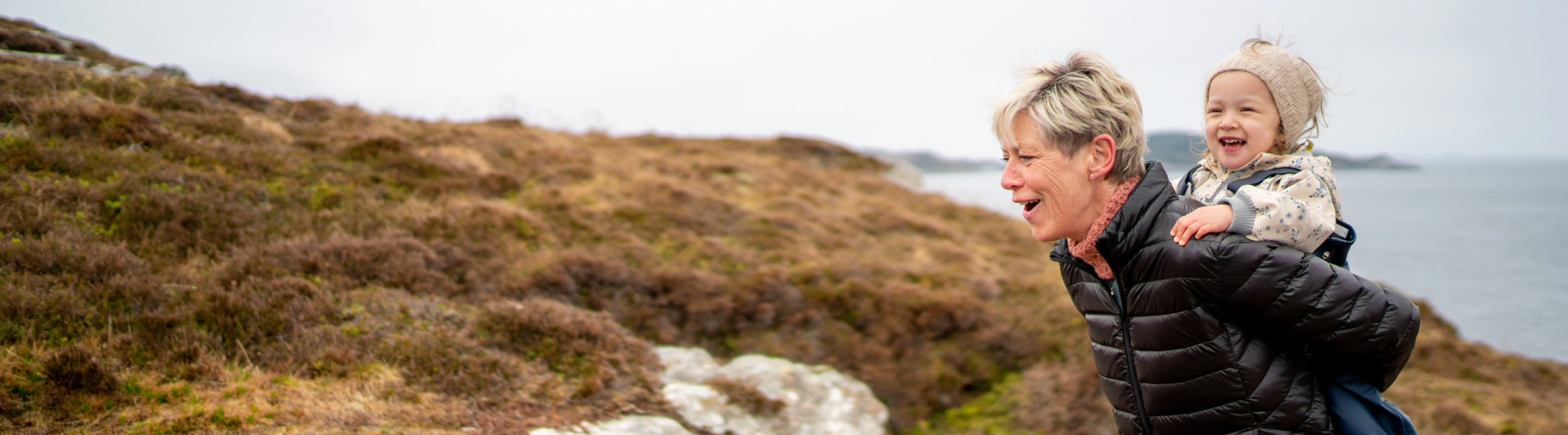 a grandmother with travel insurance for seniors, carrying her granddaughter on her back on a hike by the ocean