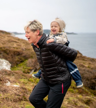 a grandmother with travel insurance for seniors, carrying her granddaughter on her back on a hike by the ocean