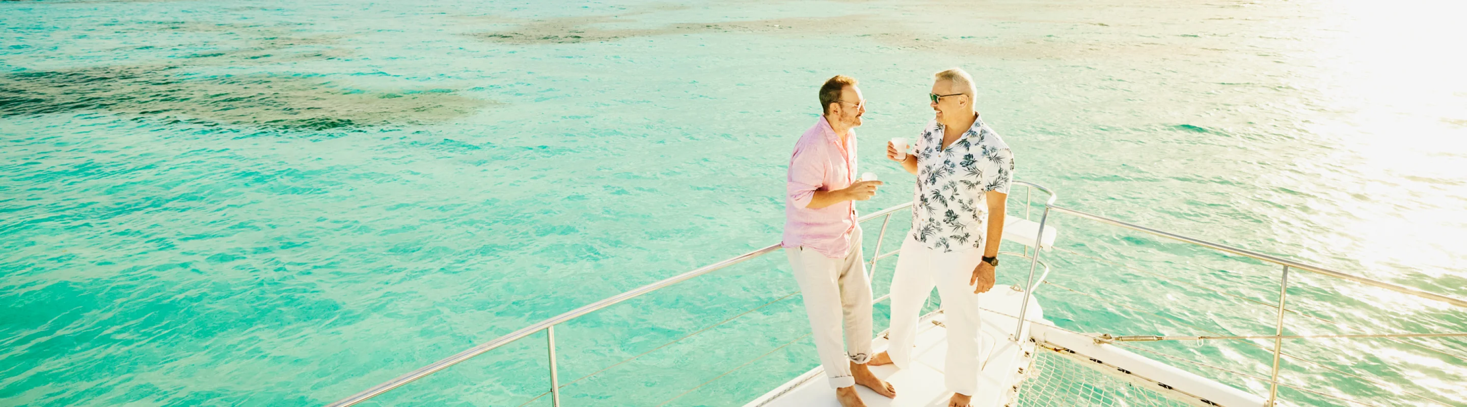 Two men with travel insurance for the Caribbean holding drinks and talking by the edge of a boat in the ocean