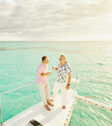 Two men with travel insurance for the Caribbean holding drinks and talking by the edge of a boat in the ocean