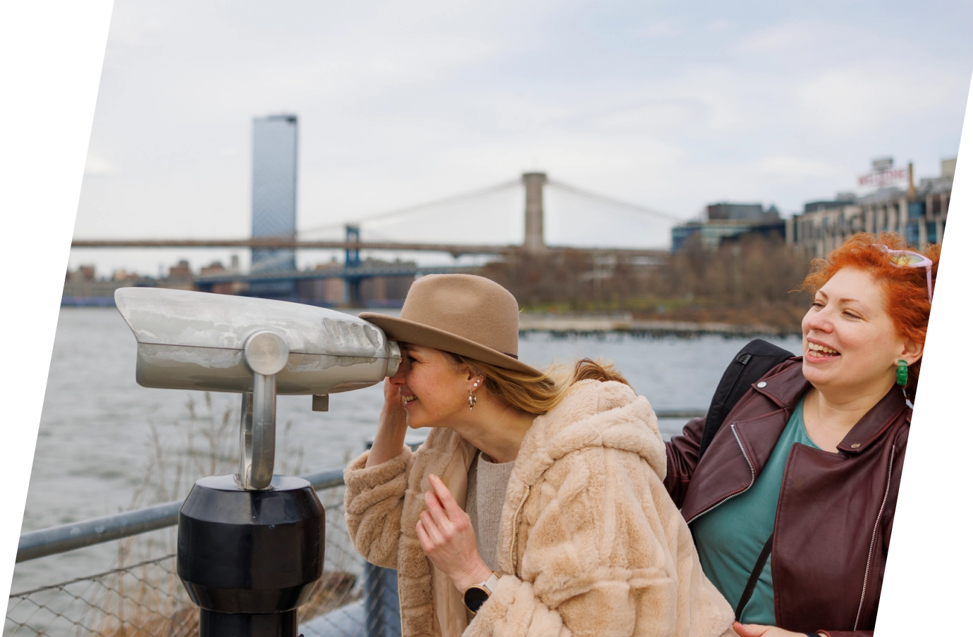 a woman with domestic travel insurance uses viewing binoculars on Brooklyn's waterfront while her friend smiles behind her