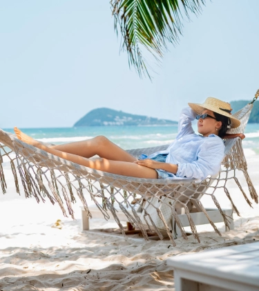 a woman with travel insurance for beach trips lies in a white hammock in between palm trees at a white-sand beach