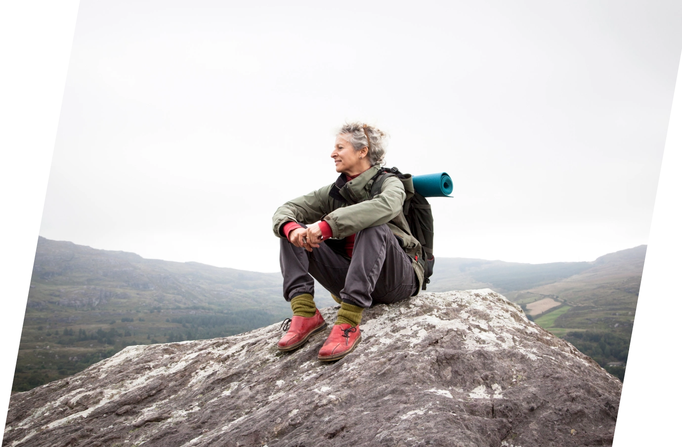 an elderly woman with baggage insurance sits on a large rock and takes a break during her hike