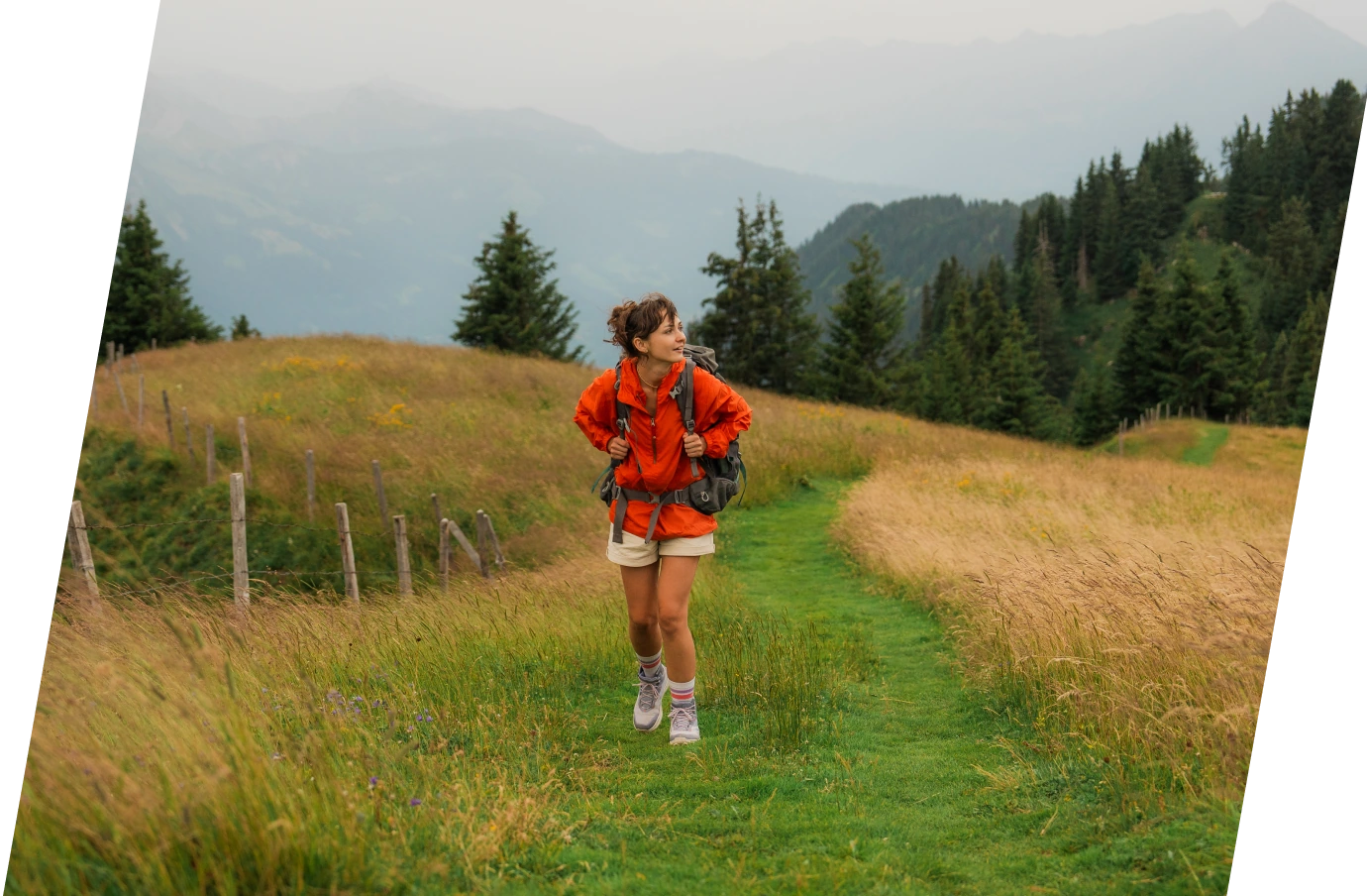 a woman with a red jacket and travel insurance for Switzerland hikes a grassy, mountain trail on vacation