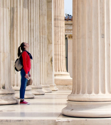 a young woman with cancel for any reason travel insurance awes at classical architecture as she stands between marble columns 