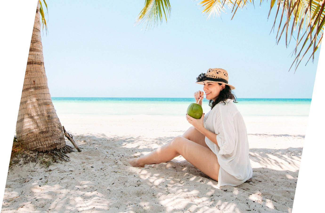 Woman holding a coconut and sitting on a white-sand beach by the shoreline with travel insurance for the Caribbean