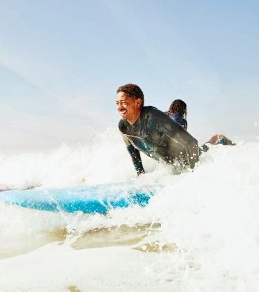 a young, mixed-race man with travel insurance for Morocco wears a wetsuit and catches a wave on his surfboard