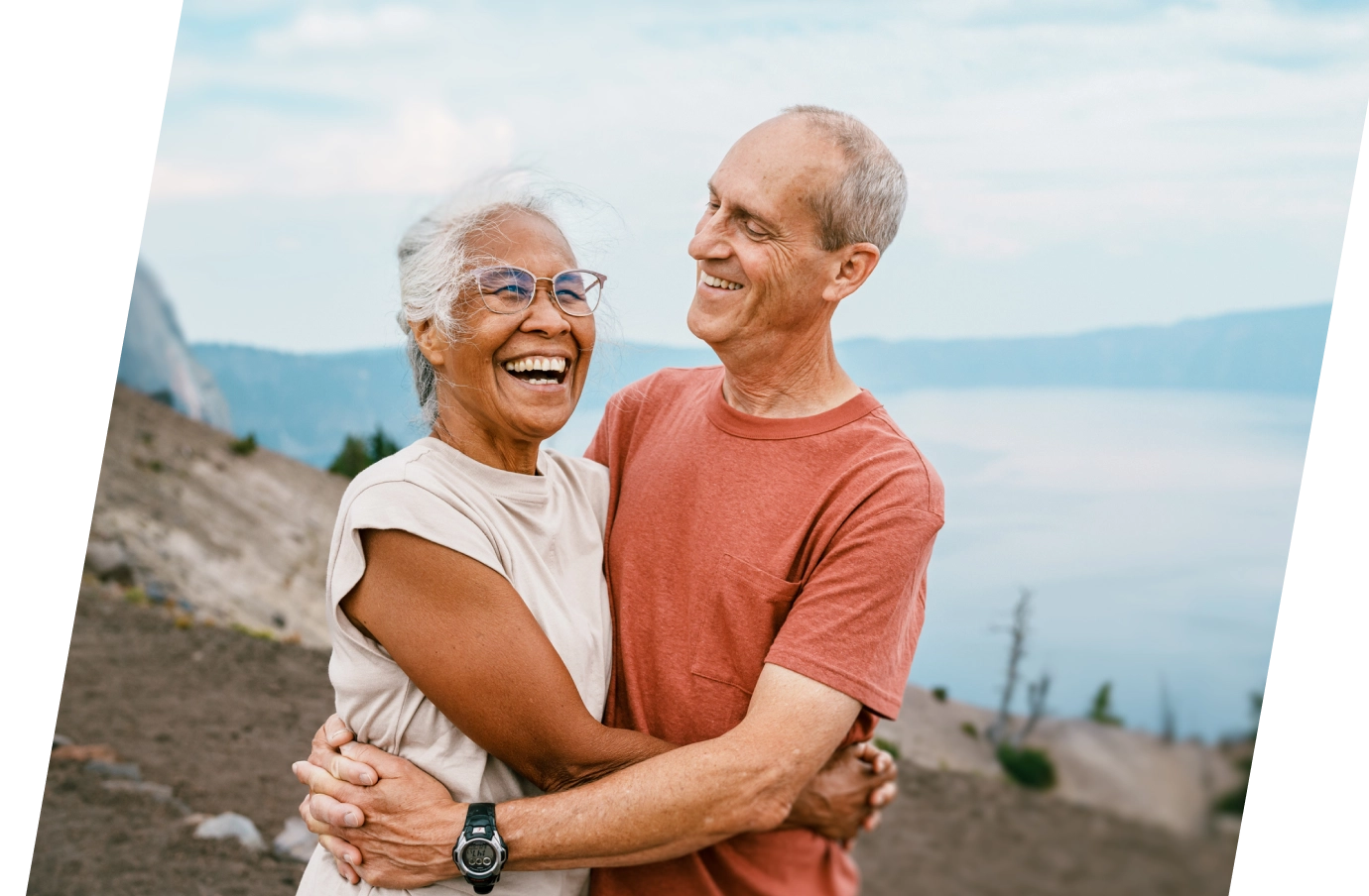 a mixed-race, elderly couple with travel insurance for nature trips embraces on a mountain near a lake 