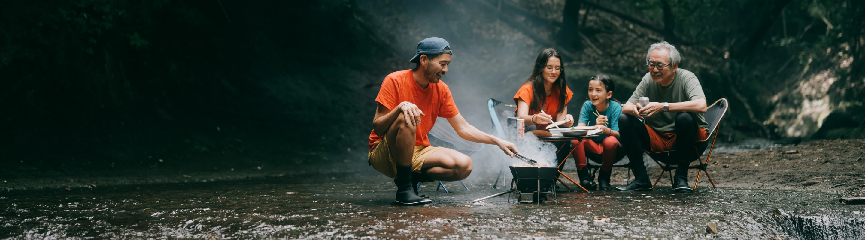 a multi-generational family with travel insurance for nature trips sits and cooks by the fire in the shallow water of a creek
