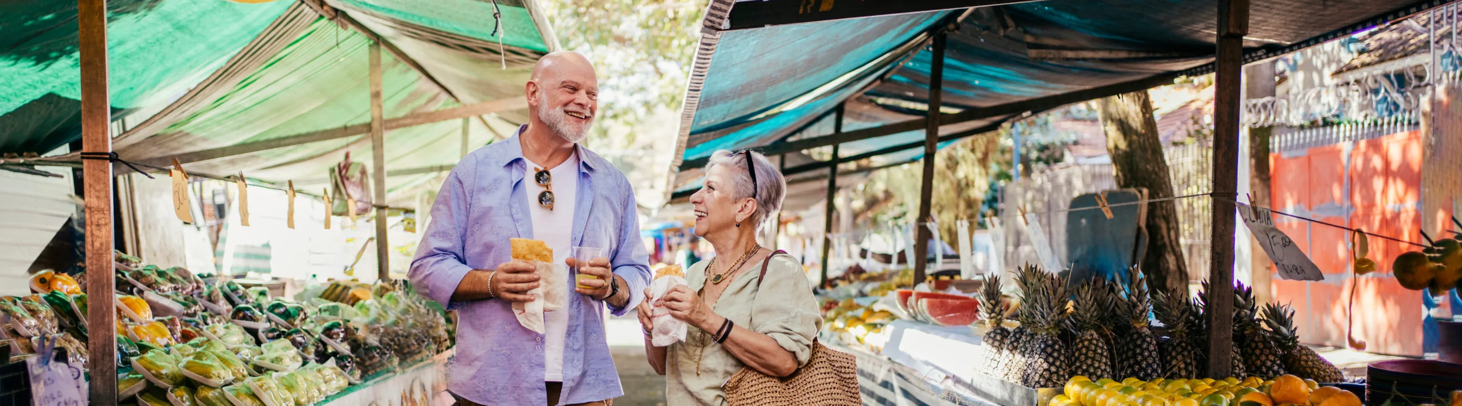 an older couple with travel insurance for culinary vacation enjoys food and drinks at a vibrant outdoor produce market 