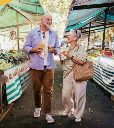 an older couple with travel insurance for culinary vacation enjoys food and drinks at a vibrant outdoor produce market 