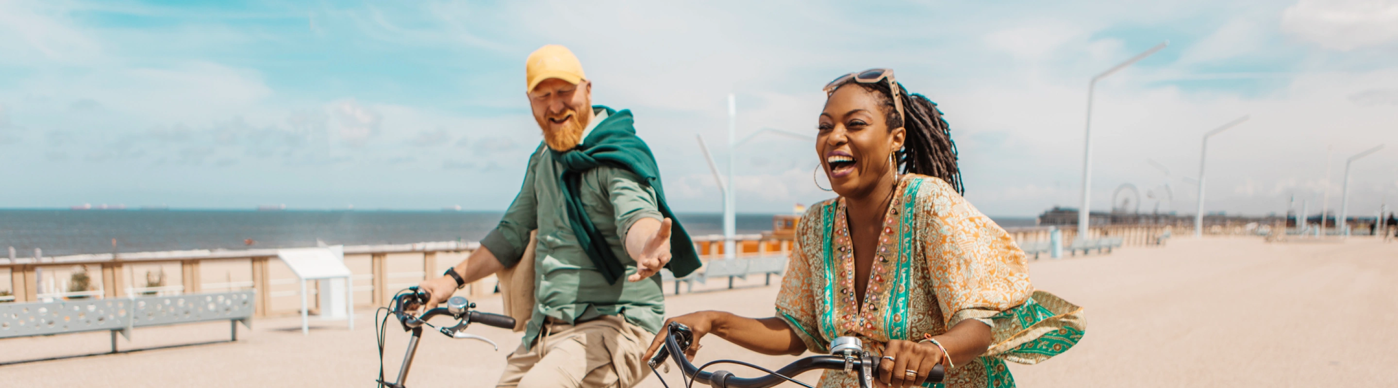 a young, mixed-raced couple with couples' travel insurance rides bikes on an oceanfront boardwalk on a sunny day