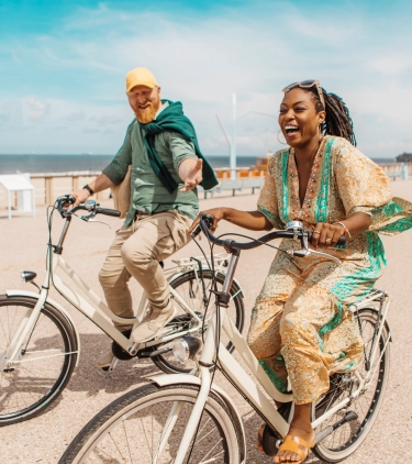 a young, mixed-raced couple with couples' travel insurance rides bikes on an oceanfront boardwalk on a sunny day