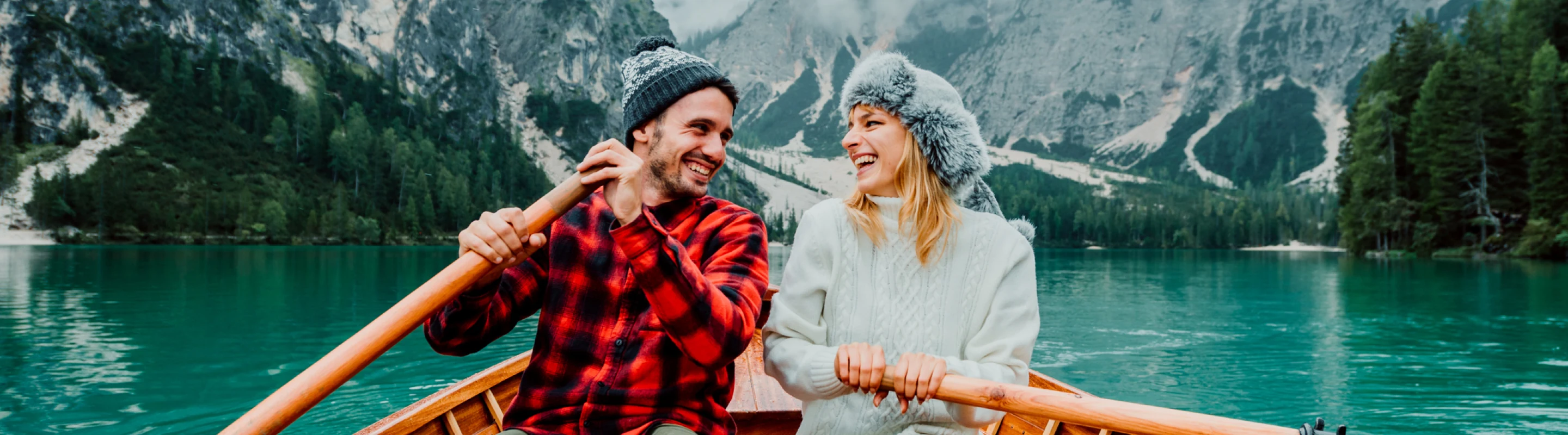a young couple wearing hats and warm clothes rows a wooden boat on a mountain lake, insured with couples' travel insurance 