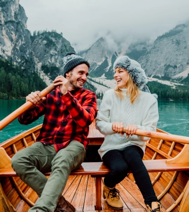 a young couple wearing hats and warm clothes rows a wooden boat on a mountain lake, insured with couples' travel insurance 